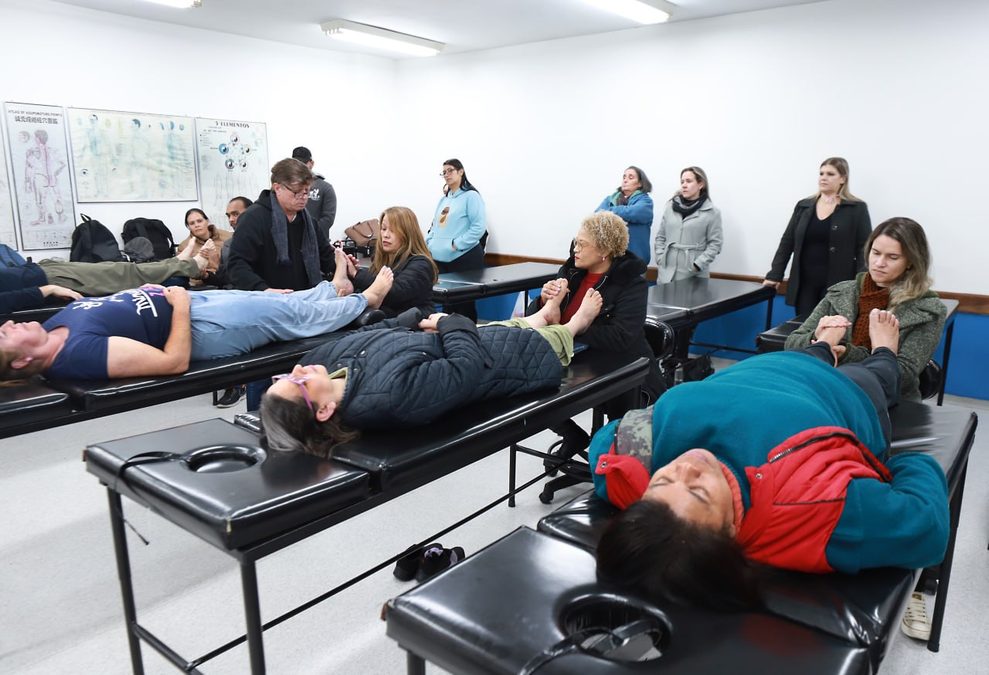 Alunos e alunas em aula prática de reflexologia com modelos convidados. Modelos deitados em macas especiais e outros alunos observando